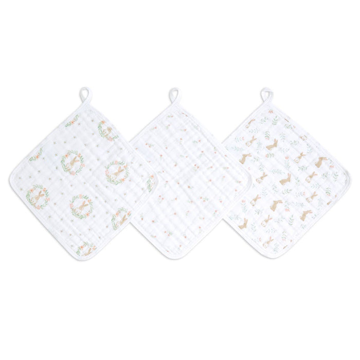The aden + anais essential cotton muslin washcloths 3 pack includes three cloths with pastel floral wreaths, small dots, and animal patterns. Ideal for delicate skin, each cloth has a loop for hanging and is displayed in a row on a flat surface.