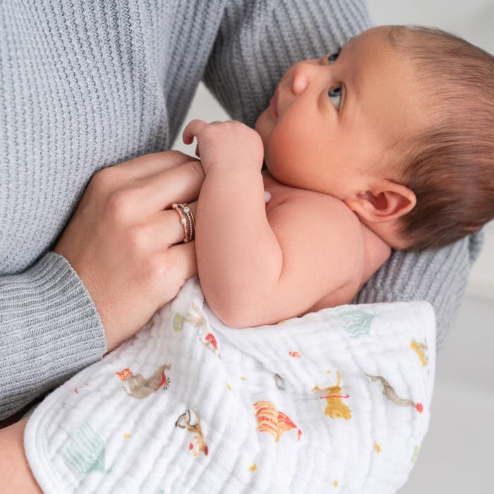 An individual wearing a gray sweater holds a baby wrapped in aden + anaiss essential cotton muslin washcloth, beautifully patterned in white, while the infant looks up at them.