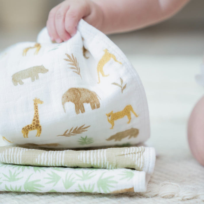 A baby holds an aden + anais essential cotton muslin washcloth featuring a giraffe and elephant. Nearby, two more absorbent, green-patterned layers are neatly folded on a light surface, perfect for delicate skin.