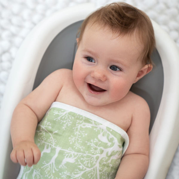 A smiling baby with short brown hair is cozily wrapped in a green aden + anais essential cotton muslin washcloth featuring a white tree pattern, nestled in a white baby bathtub. The soft fabric gently touches the babys delicate skin against a light, textured background.