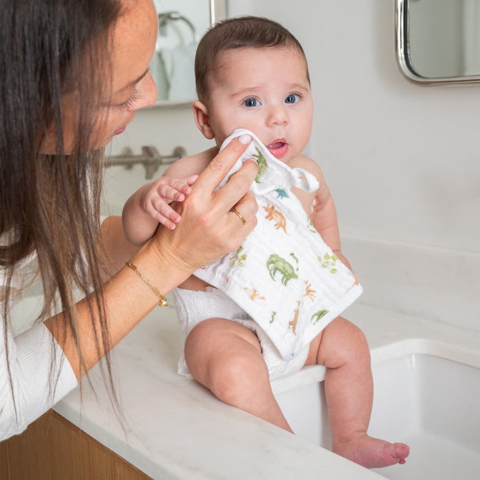 An adult gently wipes a babys delicate skin with aden + anais essential cotton muslin washcloths in a bright bathroom, where the baby sits on the counter wearing a diaper and an animal-print bib. The scene features a mirror in the background.