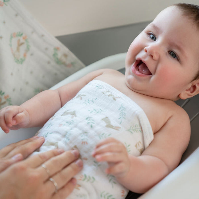 A baby smiles on a changing table, wrapped in a white blanket with animal designs. An adults hand gently touches the babys stomach, mindful of their delicate skin, while the soft patterned fabric complements the serene moment. Featured: aden + anais essential cotton muslin washcloths 3 pack.