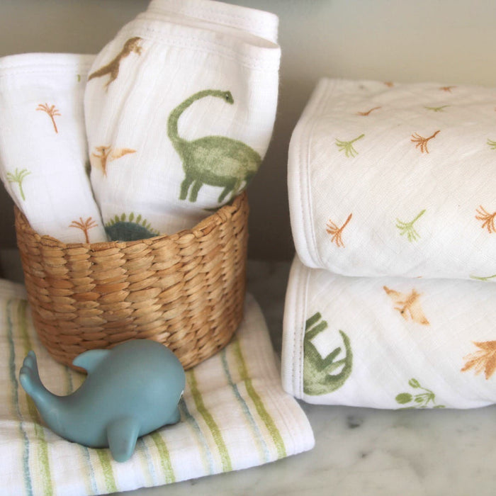 A woven basket holds a 3-pack of aden + anais essential cotton muslin washcloths, adorned with dinosaur prints; nearby, a blue whale-shaped toy rests on a striped cloth—perfect for delicate skin in the cozy nursery or baby changing area.