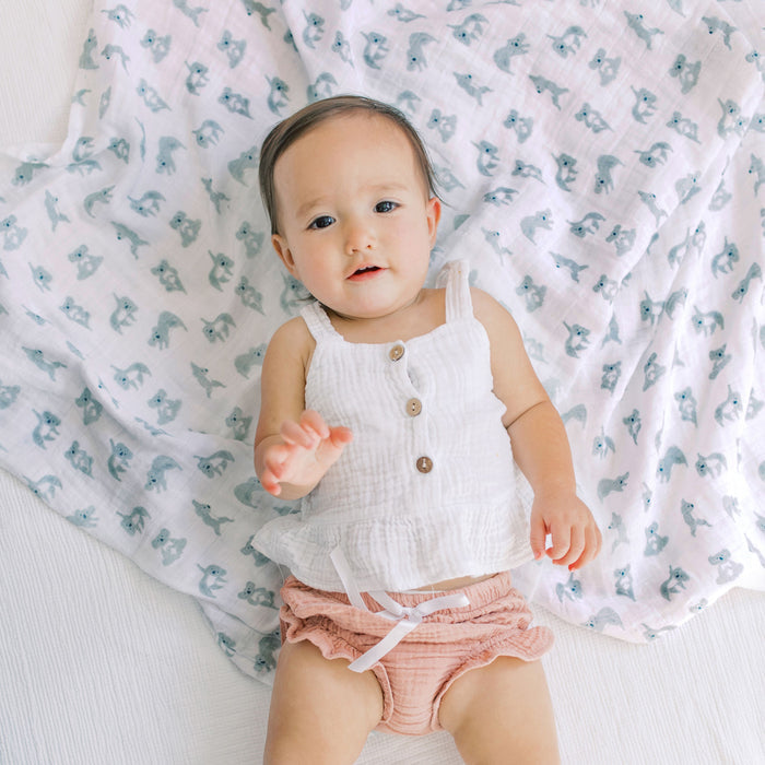 A baby in a white sleeveless top and pink shorts lies on an animal-print blanket. With curiosity, the baby reaches out, their aden + anais essentials cotton muslin bandana bib from the 2 pack adding style and comfort to the cute scene.