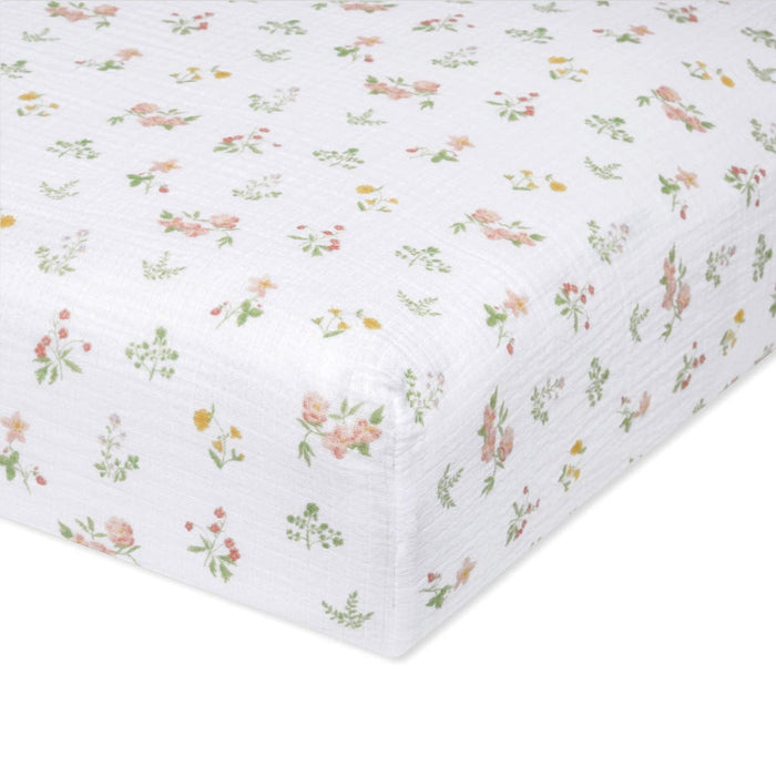 Close-up of the aden + anais essentials cotton muslin crib sheet, featuring a white, rectangular design with a floral pattern of small pink and yellow flowers and green leaves. The fabric looks soft and textured, similar to delicate nursery prints.