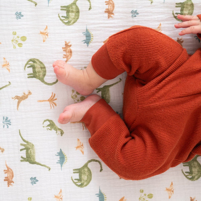 A baby in a rust-colored onesie is snuggled on the Breathable Nursery Bundle by HALO, lying on a white blanket adorned with green, orange, and blue dinosaur patterns.