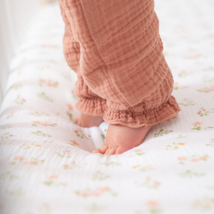A babys feet are on a soft, floral bedspread, wearing brown pants with ruffled cuffs. Nearby, the HALO Breathable Nursery Bundles Muslin Crib Sheet peeks out, adding to the cozy and gentle vibe.