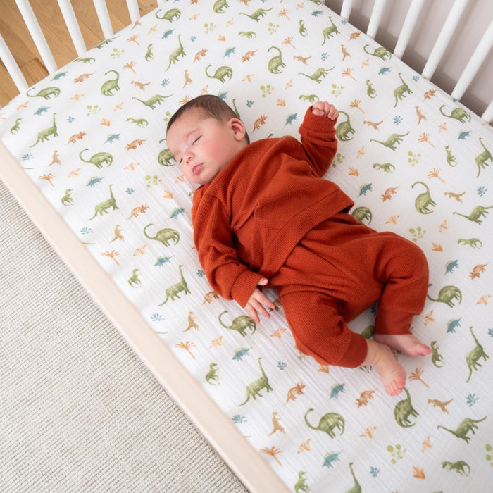 A baby sleeps peacefully in a crib, wearing a rust-colored outfit, on aden + anais essentials cotton muslin crib sheets. These feature a playful dinosaur pattern with colorful dinosaurs scattered across the fabric, perfectly sized for standard crib mattresses and ensuring a breathable nights rest.