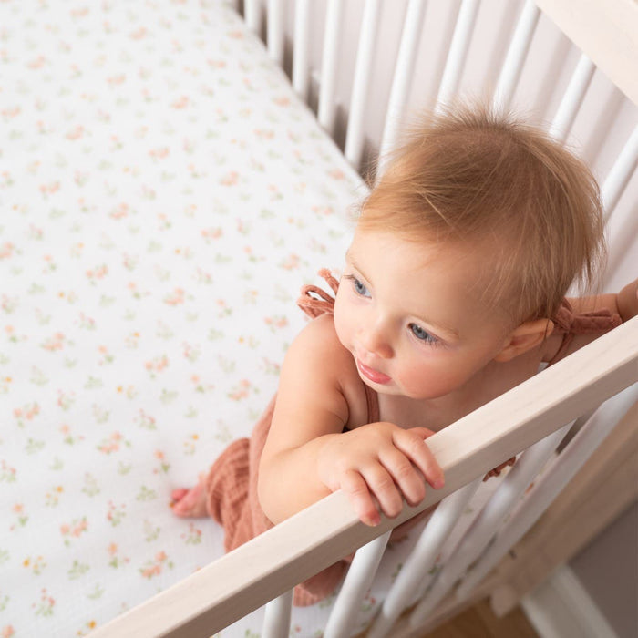 A baby stands in a white crib with an essentials cotton muslin crib sheet by aden + anais. Wearing a pink dress, the baby looks curiously over the side. Bright lighting highlights the babys light hair and the cribs clean design.