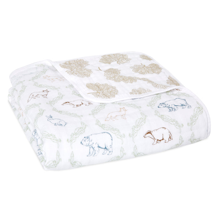 The aden + anais essentials cotton muslin dream blanket is a folded, soft cotton blanket with outlined illustrations of bears, rabbits, and raccoons in muted colors, featuring floral and leaf patterns in a light pastel palette.