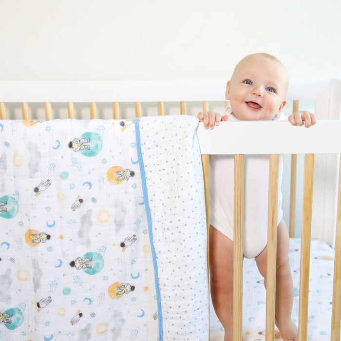 A smiling baby stands in a wooden crib, holding the rail, with an aden + anais essentials cotton muslin dream blanket featuring playful animal and moon designs draped over the side, adding charm to the scene.