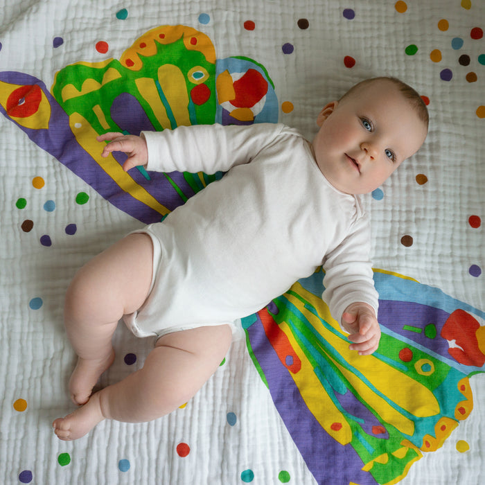 A baby in a white onesie lies on the aden + anais essentials cotton muslin dream blanket, posed so the butterfly design on the soft blanket gives the appearance of vibrant wings behind them.