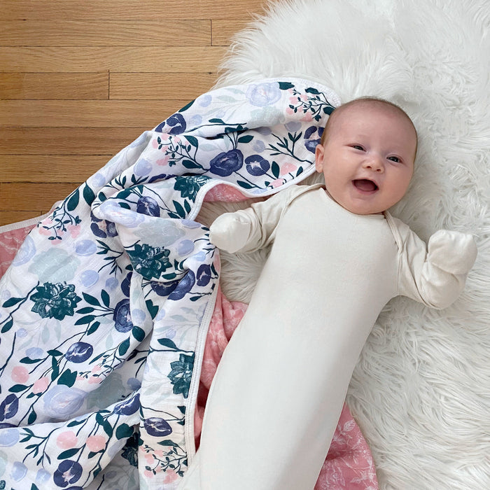 A baby in a cream sleep sack is smiling on a fluffy white rug, with an aden + anais essentials cotton muslin dream blanket in a pink and blue floral pattern on the wooden floor beside them.