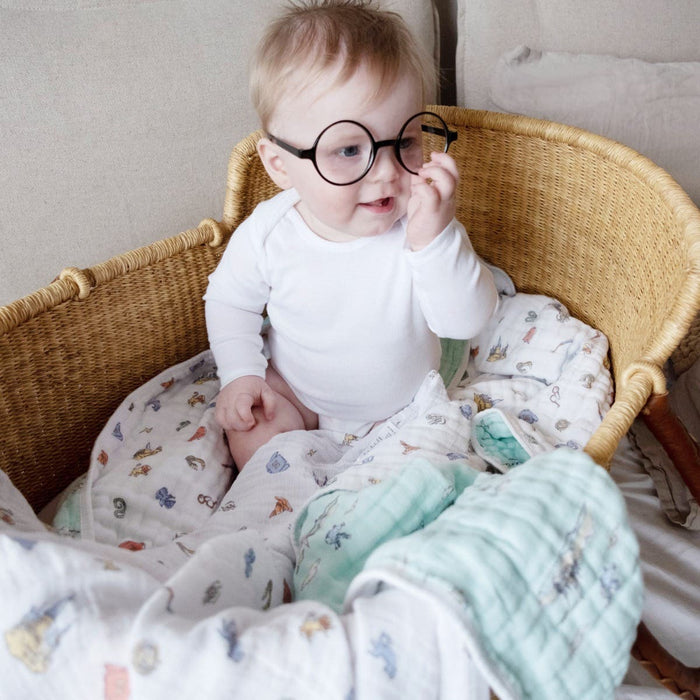 A baby in oversized round glasses sits in a woven basket lined with an aden + anais essentials cotton muslin dream blanket. Dressed in white, the little one touches the glasses, appearing curious and playful amidst soft, lightweight comfort.