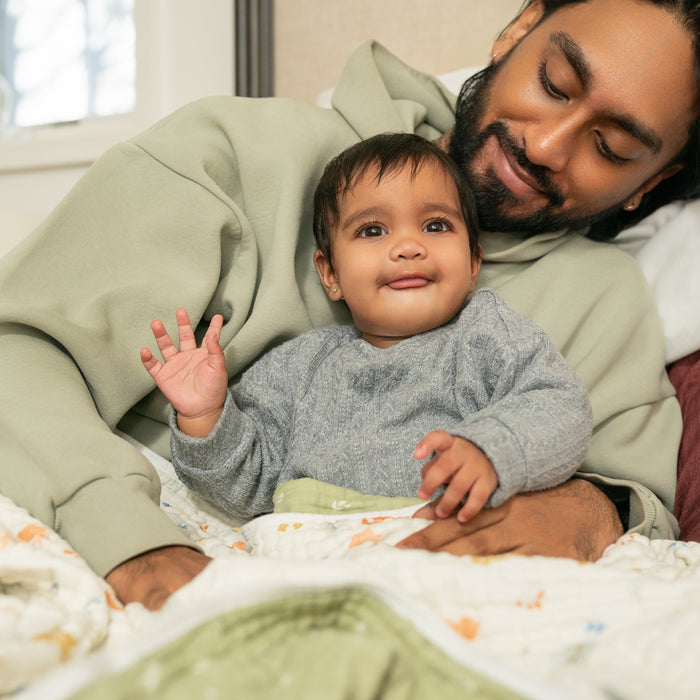 A man in a green hoodie lies on a bed, embracing a smiling baby in a gray sweater. The baby waves and sticks out their tongue as they both enjoy the warmth of aden + anais essentials cotton muslin dream blanket, adding comfort to the tender moment.