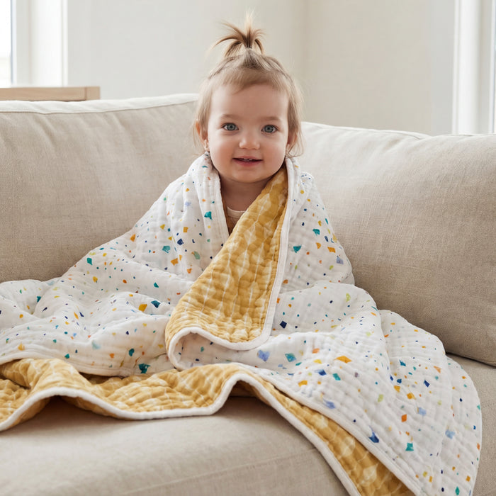 A smiling toddler with light brown hair sits on a beige sofa, wrapped in an aden + anais essentials cotton muslin dream blanket featuring colorful speckles and a yellow underside, as bright natural light fills the room.