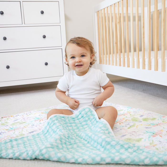 A baby with curly hair sits happily on an aden + anais essentials cotton muslin dream blanket in a nursery, wearing a white onesie. Behind the baby is a crib with wooden rails and a white dresser with black knobs.