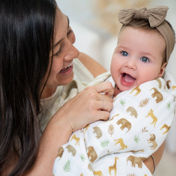 A woman smiles warmly at a baby wrapped in an aden + anais essentials cotton muslin dream blanket featuring playful animal prints. The brown headband-wearing baby grins broadly, creating a bright and cheerful scene.
