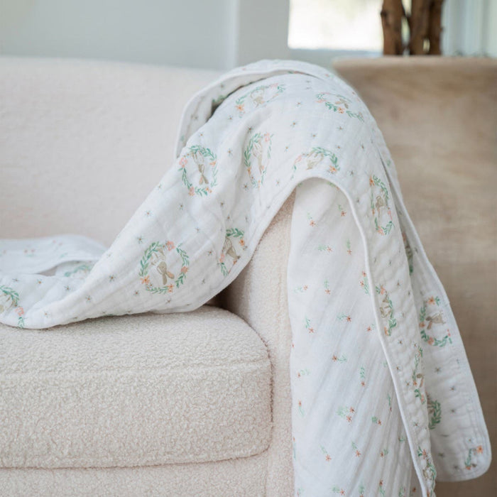 A cozy cloud of tranquility, this white chair is adorned with an aden + anais essentials cotton muslin dream blanket draped over the armrest. The blankets small, colorful floral designs and circular motifs add charm to the serene room setting.