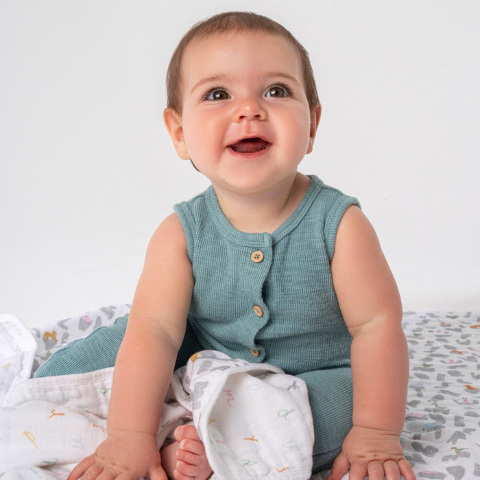 A happy baby sits on a soft surface, wearing a sleeveless, buttoned green outfit and is wrapped in an absorbent aden + anais essentials cotton muslin dream blanket. The background is neutral light gray.
