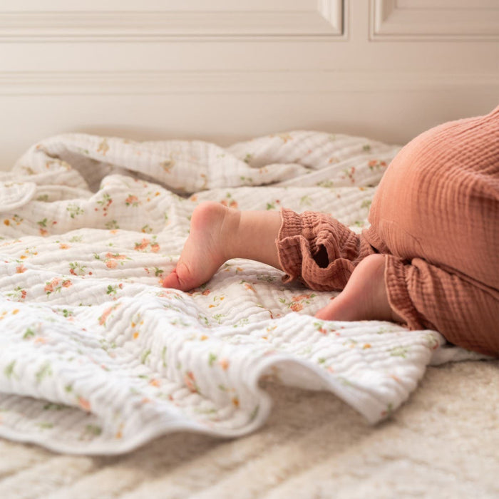 A baby lies on an aden + anais essentials cotton muslin dream blanket, their feet peeking from rust-colored textured pants in a cozy, warm setting illuminated against a white wall.