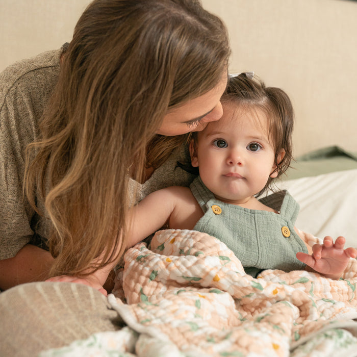 A woman with long hair kisses a toddler on the head. The toddler, in a green outfit, snuggles while wrapped in an aden + anais essentials cotton muslin dream blanket, looking at the camera on a light-colored bed.