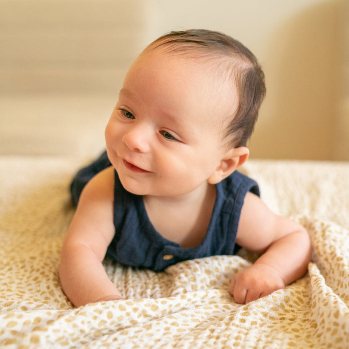A baby in a blue sleeveless outfit lies on the aden + anais essentials cotton muslin dream blanket, enjoying tummy time with a happy smile. The softly blurred background emphasizes this delightful moment on the cozy, patterned blanket.