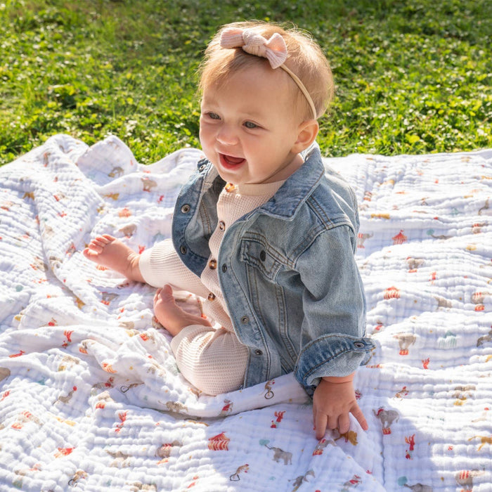 A smiling baby sits outdoors on an essentials cotton muslin dream blanket by aden + anais, wearing a denim jacket, a light outfit, and a pink bow headband. The lush green grass background creates a charming and serene setting.