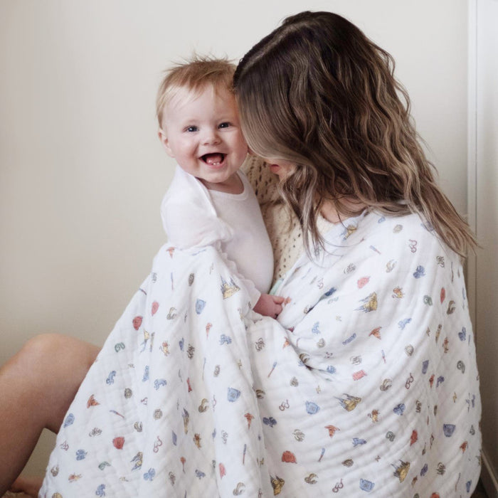A smiling baby in a white onesie is snugly held by an adult with wavy hair, wrapped in aden + anais essentials cotton muslin dream blankets. They sit contentedly on the floor against a plain wall, with the baby happily looking at the camera.