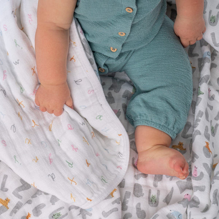 A baby in green clothing sits on a patterned blanket, holding the aden + anais essentials cotton muslin dream blanket adorned with colorful letters. The absorbent lightweight fabric and the babys visible feet create a cozy and gentle atmosphere.