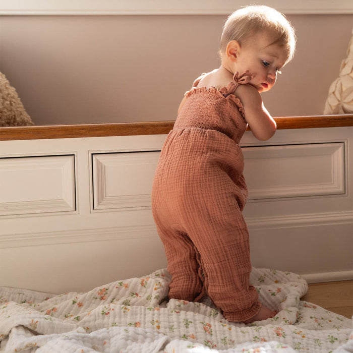 A baby in a soft, brown romper stands while holding onto a wooden bench in a softly lit room with white paneled walls. On the floor are floral-patterned aden + anais essentials cotton muslin dream blankets. The baby looks to the side with a curious expression.