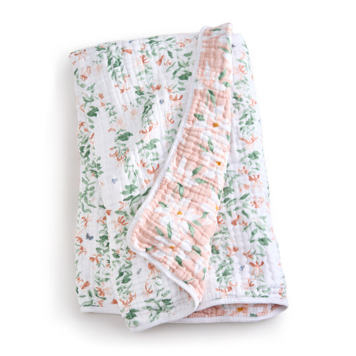 The aden + anais essentials cotton muslin dream blanket is a folded, quilted blanket featuring coral floral and green leaf prints on a white base. It promises cozy snuggles with its white-trimmed edges and soft blush pink underside.