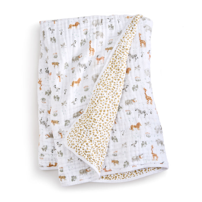 The aden + anais essentials cotton muslin dream blanket is a cozy, white baby blanket featuring a safari animal print with giraffes, elephants, and lions. The reverse side has beige polka dots. Its perfect for tummy time or cuddling and is displayed partially folded.