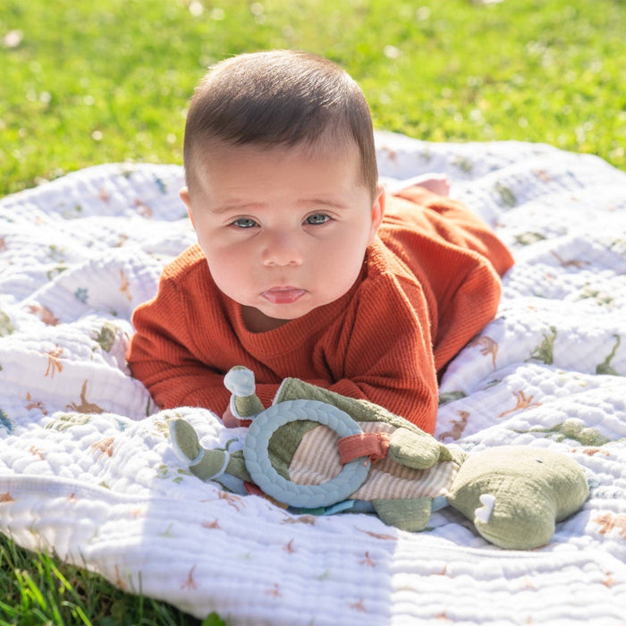 A baby in an orange outfit lies on the soft grass, cradled by an aden + anais essentials cotton muslin dream blanket, holding a toy. Sunlight gently bathes the scene, creating a warm and cheerful atmosphere.