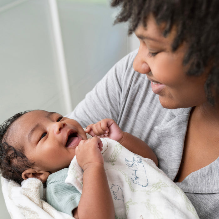 A smiling caregiver gazes lovingly at an infant wrapped in an aden + anais essentials cotton muslin dream blanket. The baby, wearing a light-colored onesie, looks up joyfully as the background softly blurs.