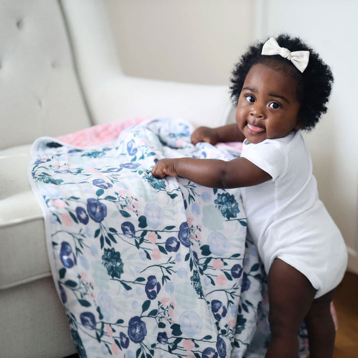A baby in a white onesie and bow stands holding an aden + anais essentials cotton muslin dream blanket with a colorful, floral pattern draped over an armchair. The soft lighting enhances the cozy atmosphere, perfect for snuggling with these warm, absorbent blankets.
