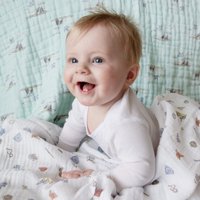 A smiling, light-haired baby with blue eyes sits on a cushioned surface, wearing a white outfit. The background features aden + anais essentials cotton muslin dream blanket adorned with cozy clouds and small, playful patterns in soft pastel colors.