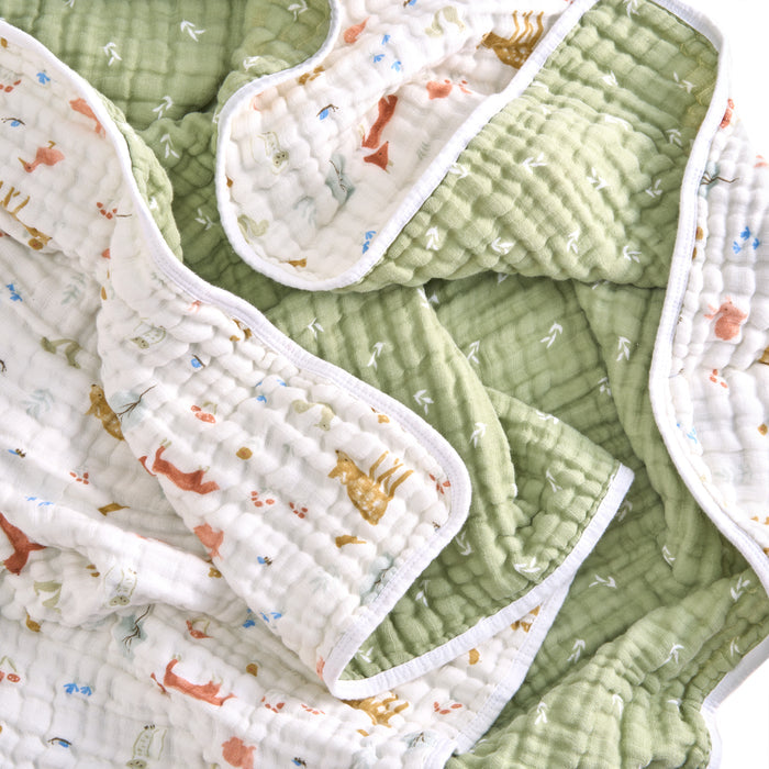 The aden + anais essentials cotton muslin dream blanket is a cozy, quilted baby blanket with a green side and a white side featuring cartoon giraffes and foxes. Made from soft, absorbent cotton muslin, it is slightly folded to display both sides and their contrasting patterns.