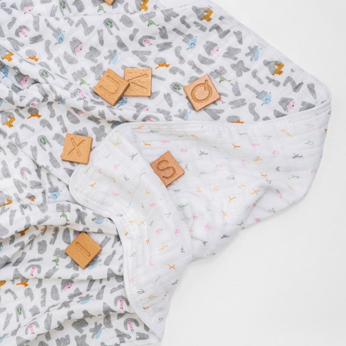 The aden + anais essentials cotton muslin dream blanket, adorned with pastel giraffes and elephants, is showcased alongside wooden letter blocks like X, U, C, and S. Perfect for babies, this soft blanket provides comfort in gentle hues.
