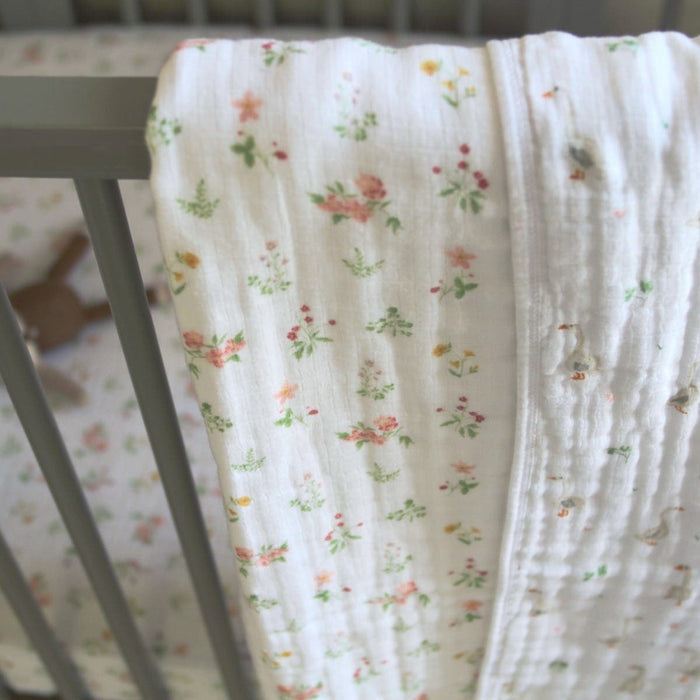 An essentials cotton muslin dream blanket by aden + anais, in soft pastel floral, drapes over a gray crib rail like a cozy cloud. The bedding matches the floral theme, and a plush toy is nestled inside, crafting a serene ambiance.