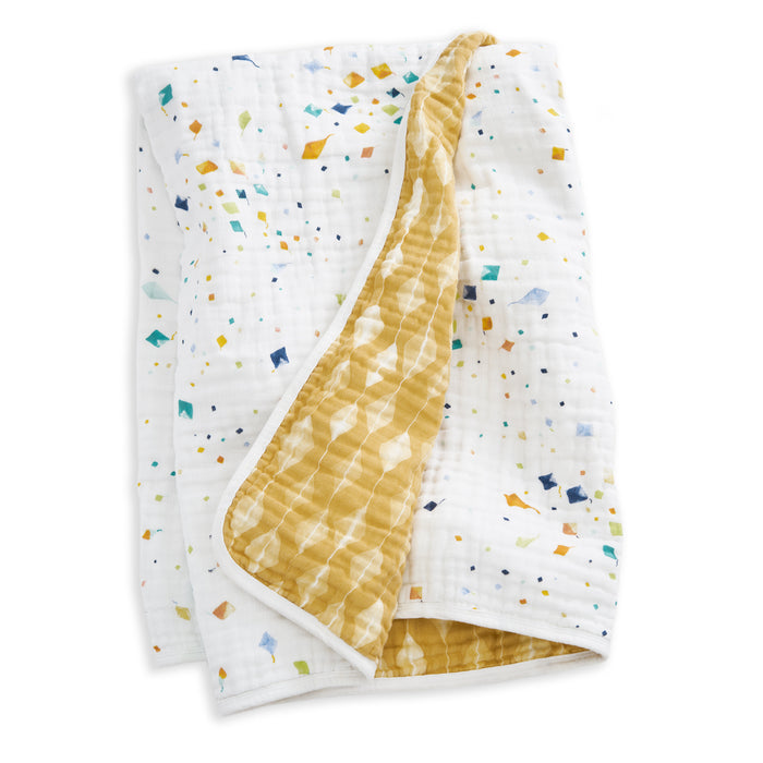 The aden + anais essentials cotton muslin dream blanket features a white side with colorful geometric shapes and a yellow reverse with a white abstract pattern, crafted from gentle cotton muslin for cozy comfort.