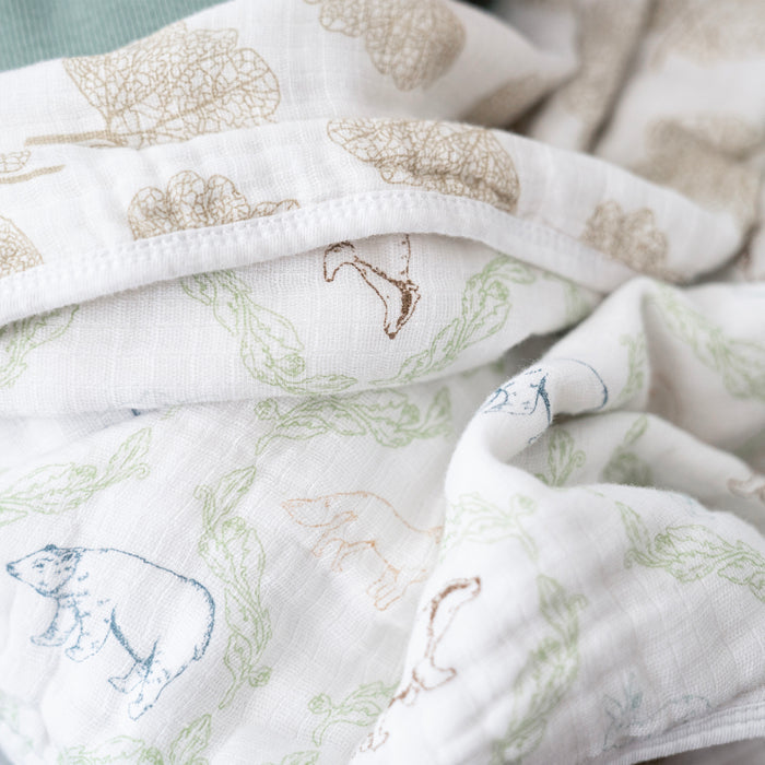 A close-up of the aden + anais essentials cotton muslin dream blanket showcases its soft, absorbent texture featuring illustrated animal patterns with bears in blue and beige and green leafy accents, slightly crumpled to highlight the detailed design.