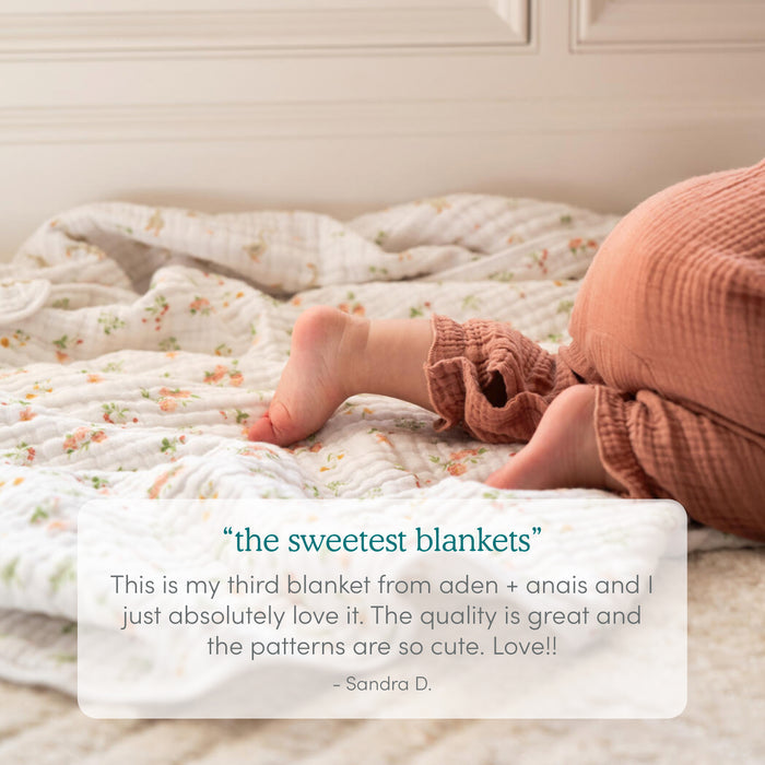 A baby in ribbed brown pants sits on a Disney™ Lilo and Stitch essentials cotton muslin dream blanket by aden + anais, with a customer review praising the newborn blanket’s quality and cute patterns shown in a white box at the bottom.