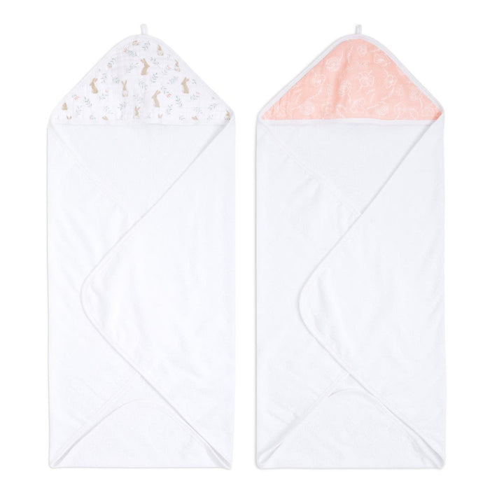 The aden + anais essentials cotton muslin hooded towels 2-pack includes a towel with white terry fabric and a multicolored animal print hood on the left, and another with a white base featuring a peach-colored hood with a subtle floral pattern on the right.