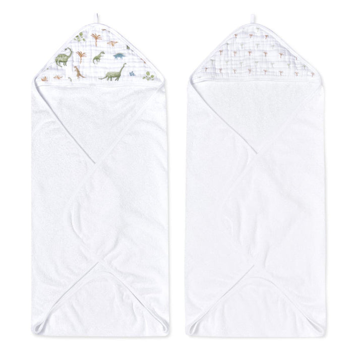 Two white aden + anais essentials cotton muslin hooded towels are shown. The left towel has a dinosaur and palm tree hood pattern, and the right features a subtle small trees pattern. Both boast a triangular hood corner design.