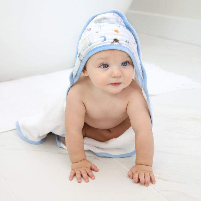 A baby crawls on a white tiled floor, wrapped in an aden + anais essentials cotton muslin hooded towel. The towels playful patterns and blue trim create charm as the absorbent fabric embraces the curious little one gazing ahead.