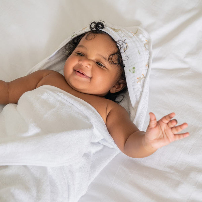 An infant grins while lying on a bed, wrapped in an aden + anais essentials cotton muslin hooded towel. The towels hood covers the babys head, and its absorbent fabric ensures comfort as the babys arm playfully stretches out.