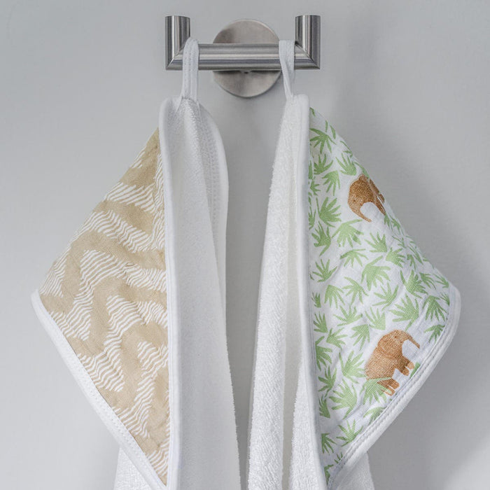 Two aden + anais essentials cotton muslin hooded towels hang on a silver hook against a white wall, one with a beige zigzag pattern and the other with green leaves and brown elephants. Both towels are super absorbent and feature white edges and loops for hanging.