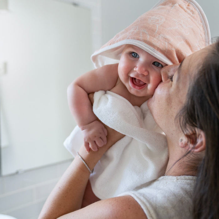 A woman kisses a laughing baby wrapped in an aden + anais essentials cotton muslin hooded towel, featuring white cotton terry and a pink hood. The background is a softly blurred indoor setting, likely a bathroom.
