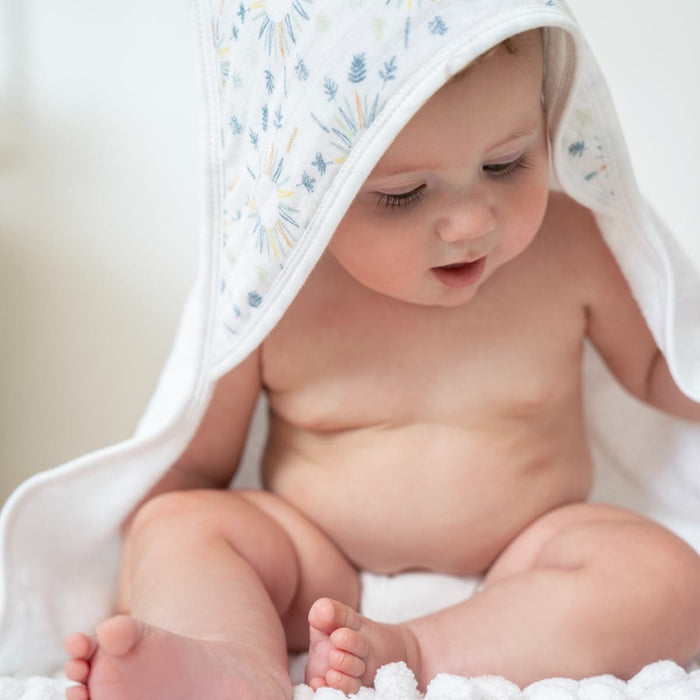 A baby sits serenely, wrapped in an aden + anais essentials cotton muslin hooded towel with a floral pattern. The soft white surface and cozy cotton terry texture in the blurred background enhance the charm of their crossed legs and delicate features.